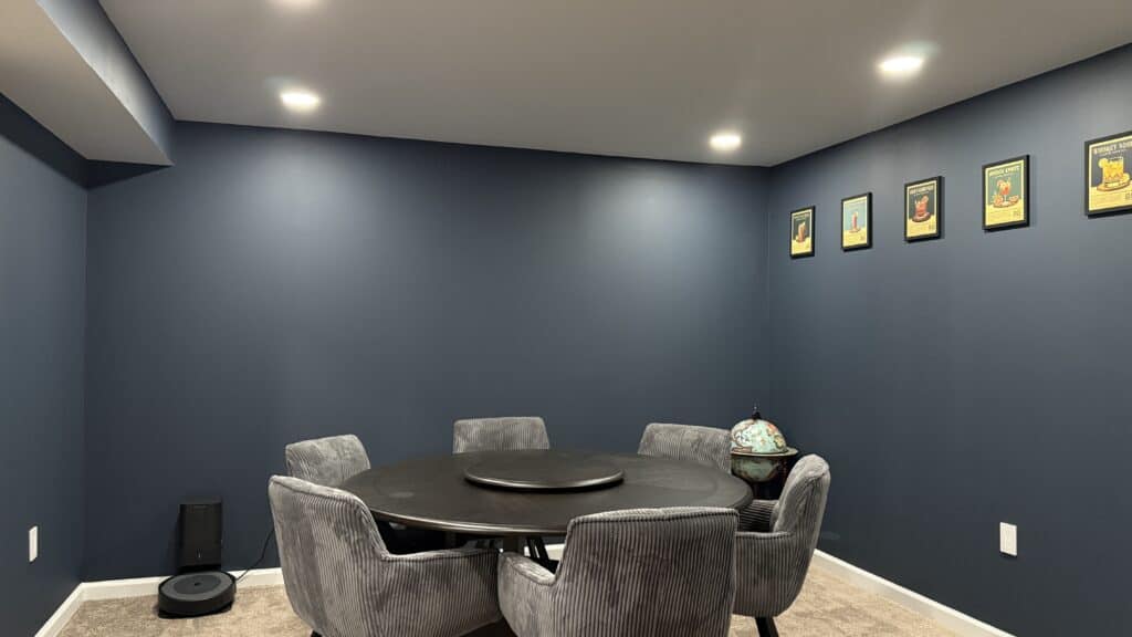 Modern basement game room with round table and six plush chairs, blue walls, and framed artwork.