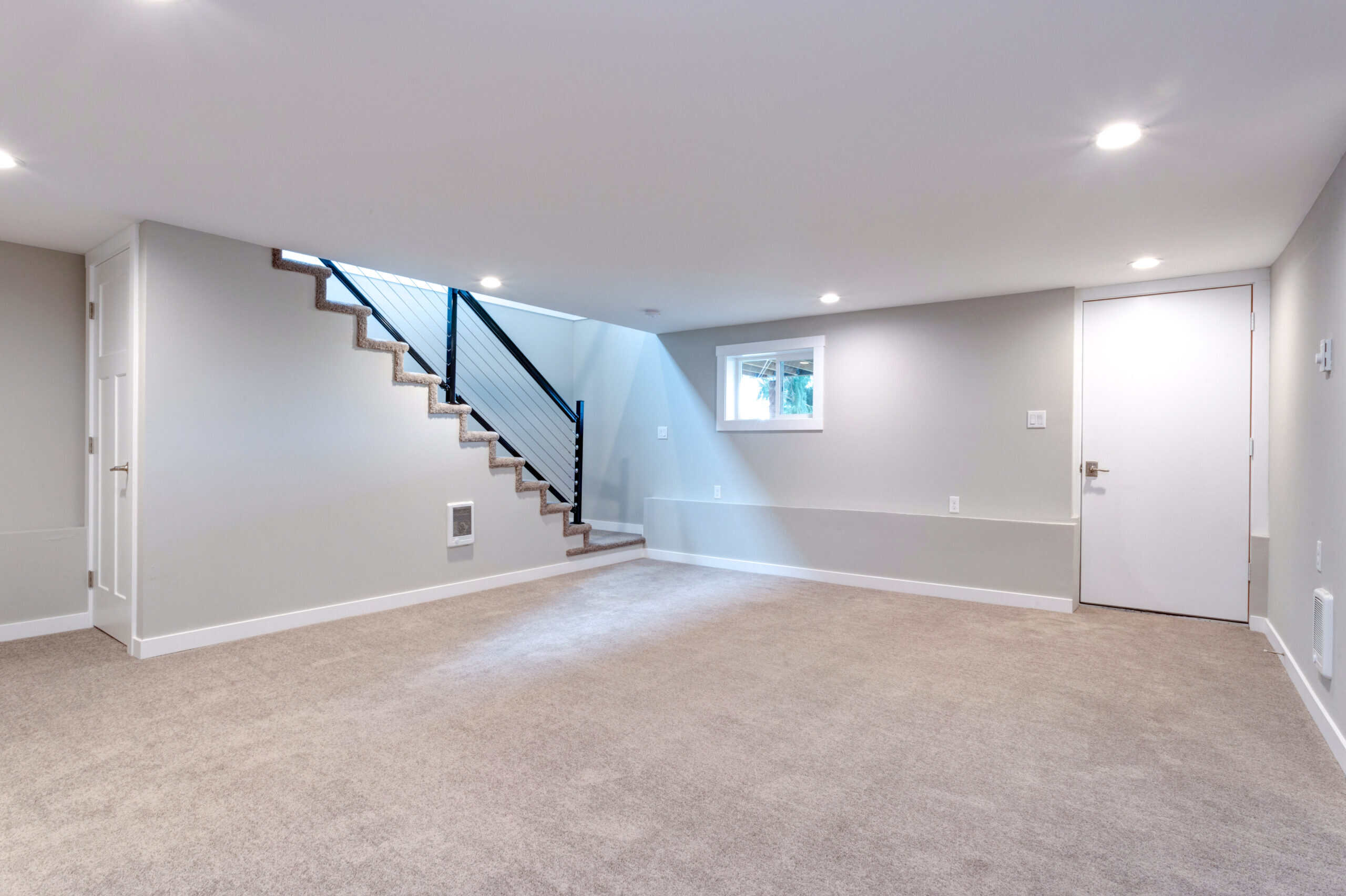 Bright, spacious basement with neutral walls, carpet flooring, and staircase with black railing.